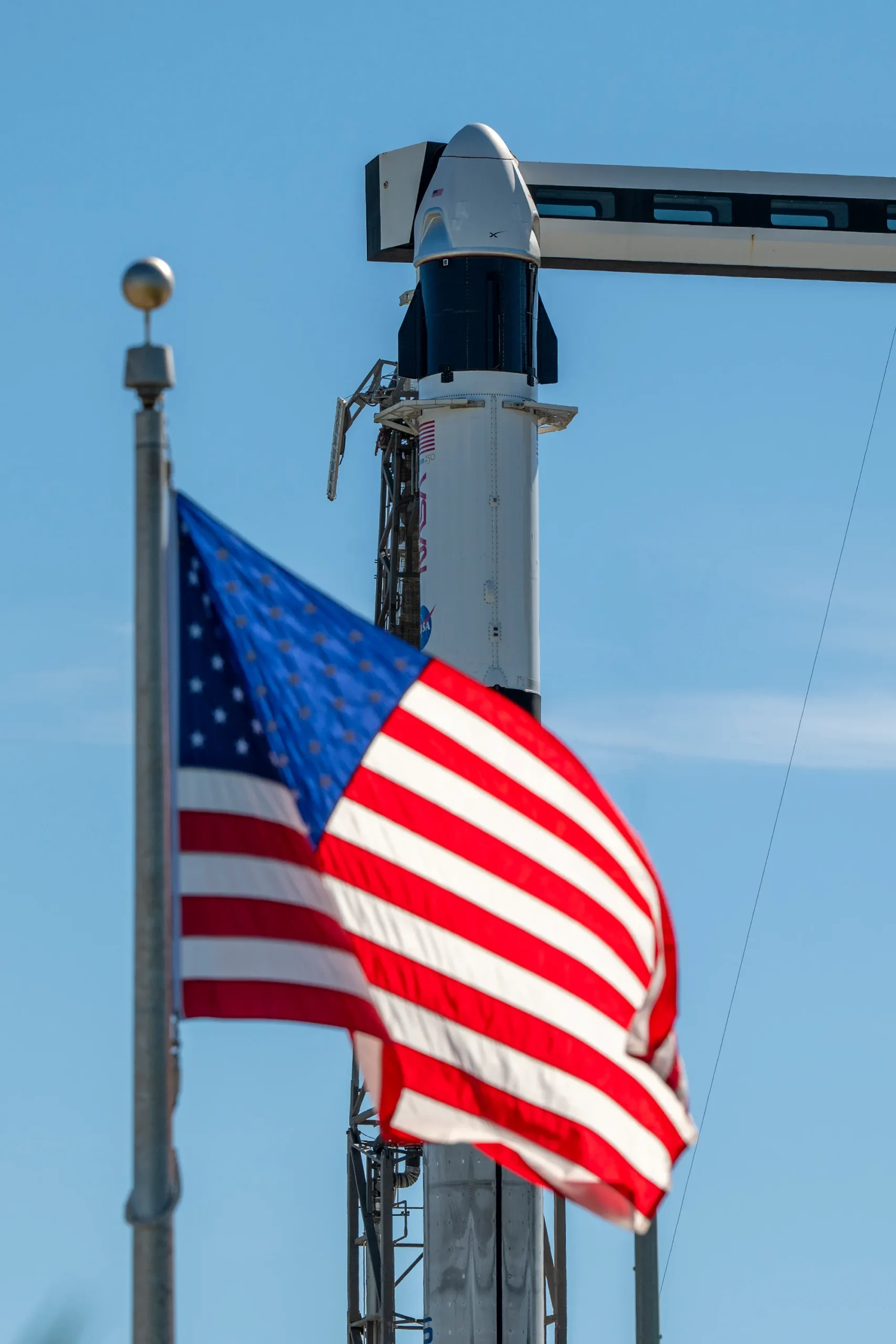A close-up view of a SpaceX Crew Dragon spacecraft mounted atop a Falcon 9 rocket on the launch pad. The capsule and trunk are connected to ground support hardware, with NASA and mission insignia visible on the rocket’s exterior against a bright blue sky. in the foreground in the American flag.