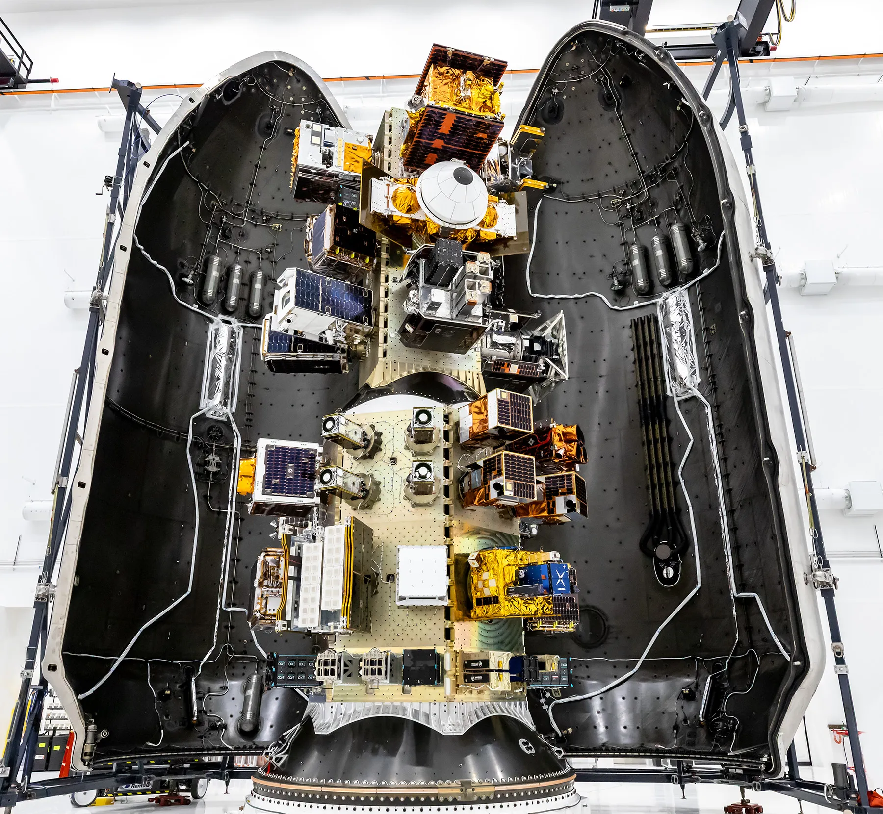Top‑down view of a SpaceX payload fairing interior showing mounted satellites and hardware arranged along the center structure, surrounded by the curved black fairing walls.